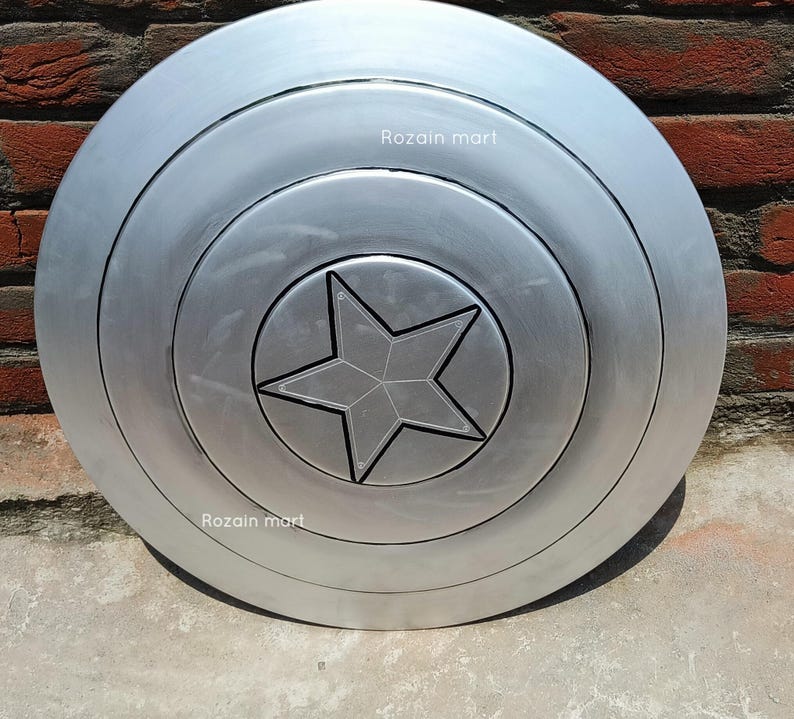 May include: A silver-colored, circular shield with a five-pointed star in the center. The shield has concentric rings and the text "Rozain mart" is visible on the surface. The shield is likely a prop or collectible.