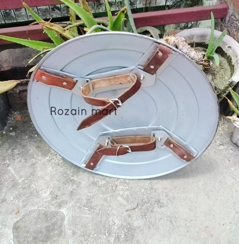 May include: A gray, circular shield with brown leather straps and buckles. The shield has a metallic appearance and features the text "Rozain mart". The straps are riveted to the shield, suggesting a durable construction.