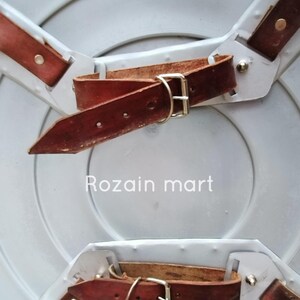 May include: A silver-colored, round shield with brown leather straps and buckles. The shield has a slightly concave surface and is designed with three leather straps attached to the back for securing it. The text "Rozain mart" is visible on the shield.