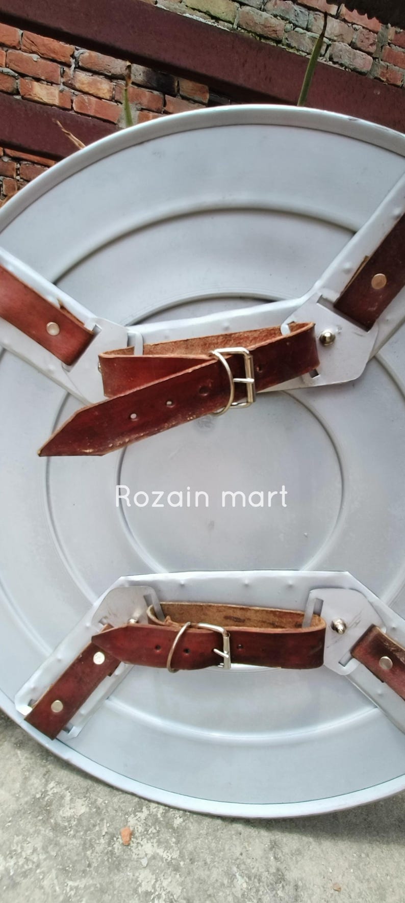 May include: A silver-colored, round shield with brown leather straps and buckles. The shield has a slightly concave surface and is designed with three leather straps attached to the back for securing it. The text "Rozain mart" is visible on the shield.