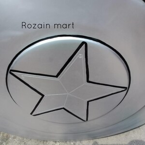May include: A silver-colored shield with a black outline of a star in the center. The shield has concentric circles and the text "Rozain mart" is visible on the top.