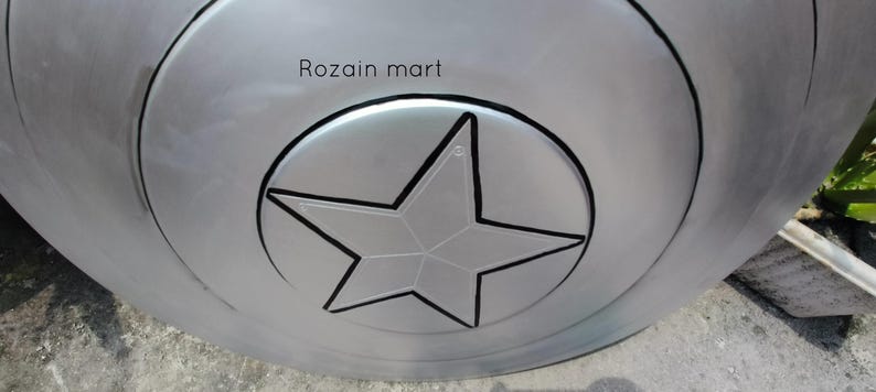May include: A silver-colored shield with a black outline of a star in the center. The shield has concentric circles and the text "Rozain mart" is visible on the top.