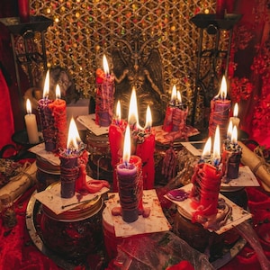 May include: A collection of red and purple candles with visible wax drips, each resting on a small square paper base atop glass jars. The candles are lit, casting a warm glow, and are arranged around a central dark statue on a red surface.