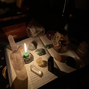 May include: An open book with handwritten text is surrounded by crystals, a lit green candle in a silver holder, and a small glass jar. The crystals are various shapes and colours, including rose quartz and clear quartz.