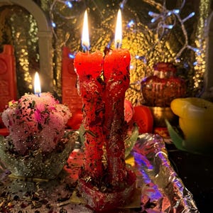 May include: Two red candles with two wicks each, burning with bright yellow flames. The candles are covered in herbs and sit on a silver tray. A smaller pink candle with a single flame is also visible, along with other decorative items.