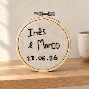 Puede incluir: Un bastidor de bordado circular de madera que contiene tela de color beige claro con el texto Inês e Marco y la fecha 23.06.26 bordados a mano en negro. El bastidor cuenta con un tornillo de tensión de metal en la parte superior.