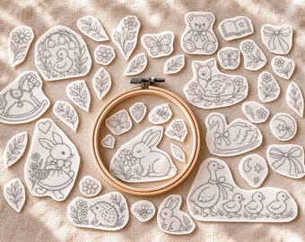 May include: A collection of white fabric patches featuring black line art embroidery designs of animals, flowers, butterflies, and bows, centered around a wooden embroidery hoop containing a rabbit patch with a flower.