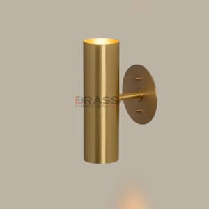 May include: A cylindrical, gold-colored wall sconce with a circular mounting plate. The sconce has a warm, inviting glow emanating from the top. The text "BRASS CHANDELIERS" is visible on the sconce.