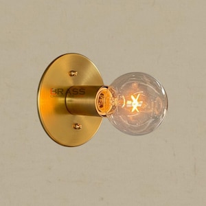 May include: A brass wall sconce featuring a round backplate and a cylindrical arm holding a clear, round light bulb. The bulb has a visible filament. The word "BRASS" is printed on the sconce.