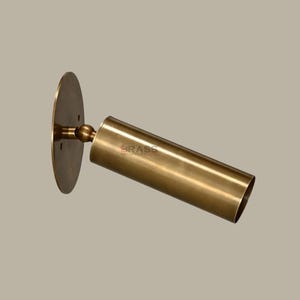 May include: A brass wall light with a cylindrical shade and a circular backplate. The fixture is angled and features the text "BRASS CHANDELIERS". The finish is a warm, metallic gold.