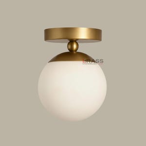 Könnte beinhalten: Eine moderne Deckenleuchte aus Messing mit einem kugelförmigen, weißen Glasschirm. Die Leuchte hat einen runden, goldfarbenen Sockel und eine passende Oberseite. Der Text "BRASS CHANDELIER LAMP" ist auf der Leuchte zu sehen.