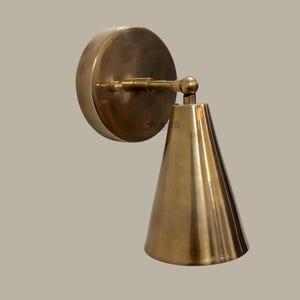 May include: A brass wall sconce with a conical shade and a circular backplate. The sconce has a warm, metallic finish and a simple, elegant design. The sconce is designed to be mounted on a wall and provides directional lighting.