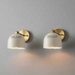 May include: Two wall-mounted sconces with a cream-colored, dome-shaped shade and a brass-colored mounting plate and arm. The sconces are lit, casting a warm glow on the wall. The text "BRASS CHANDELIER" is printed on the shade.