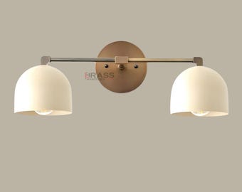 May include: A two-light wall sconce with a brass finish. The sconce features two off-white, dome-shaped shades and a horizontal arm. The fixture is mounted on a circular backplate. The text "BRASS CHANDELIER" is visible.