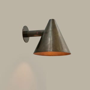 Peut inclure: Applique murale en laiton avec un abat-jour conique. L'applique a une base circulaire, un bras cylindrique et un abat-jour en forme de cône. L'intérieur de l'abat-jour dégage une lueur dorée et chaleureuse. L'applique est fixée à un mur de couleur neutre.