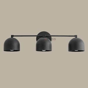May include: A black, three-light vanity fixture with a minimalist design. The fixture has three dome-shaped shades, each with a light bulb. The shades are connected by a horizontal bar and mounted on a circular base. Designed for wall mounting.
