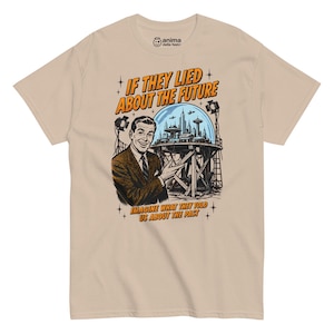 Può includere: Una t-shirt beige a maniche corte con una stampa grafica di una persona sorridente in abito che indica una città futuristica sotto una cupola di vetro. Il testo recita IF THEY LIED ABOUT THE FUTURE IMAGINE WHAT THEY TOLD US ABOUT THE PAST.