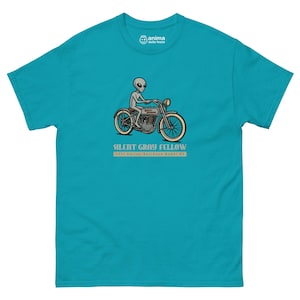 Può includere: T-shirt turchese con un alieno dei cartoni animati su una moto d'epoca. La moto è grigia con pneumatici color crema. Il testo "SILENT GRAY FELLOW" e "1937 Harley-Davidson Model 88" sono stampati sotto.