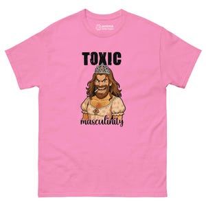 Può includere: T-shirt rosa con la scritta "TOXIC" sopra l'illustrazione di una persona barbuta con corona e vestito, con la parola "masculinity" sotto. La maglietta ha maniche corte e girocollo.
