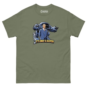 Può includere: T-shirt verde oliva con un'illustrazione a cartoni animati di un uomo in giacca e cravatta davanti a un veicolo robotico. Il testo "IT'S ONLY A GLITCH" è sotto l'immagine. La maglietta ha un girocollo e maniche corte.