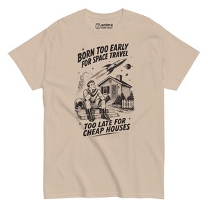 Può includere: Una t-shirt beige a maniche corte con una stampa grafica nera che raffigura una persona seduta sui gradini di una casa, un razzo nel cielo e il testo BORN TOO EARLY FOR SPACE TRAVEL e TOO LATE FOR CHEAP HOUSES, con il logo anima della festa sul collo.