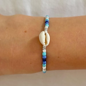 Könnte beinhalten: Ein Armband mit Perlen und einem weißen Seestern-Mittelstück. Das Armband hat eine Mischung aus blauen, silbernen und weißen Perlen. Die Seestern ist der Mittelpunkt, wobei die Perlen einen farbenfrohen Akzent setzen. Dieses Armband ist ein Schmuckstück.