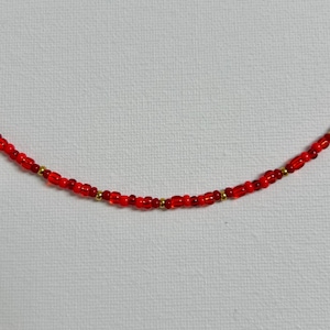 Könnte beinhalten: Eine rote Perlenkette mit kleinen, runden Perlen und einigen goldfarbenen Perlen. Die Halskette wird vor einem weißen Hintergrund präsentiert. Die Perlen sind in einem leuchtenden Rot gehalten.