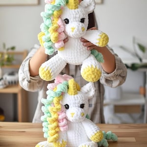 Könnte beinhalten: Zwei weiße Häkel-Einhorn-Plüschtiere mit bunten Mähnen in Rosa, Gelb und Grün. Die Einhörner haben gelbe Hufe und Hörner sowie schwarze Augen. Eines wird hochgehalten, das andere sitzt auf einer Holzoberfläche.