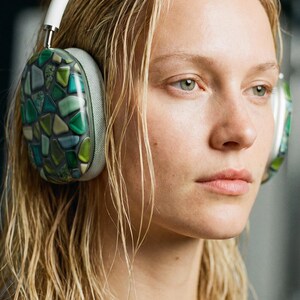 Könnte beinhalten: Weiße und silberne Kopfhörer mit Ohrmuscheln, die mit einem Mosaik aus grünen, blauen und beigen geometrischen Formen verziert sind. Die Kopfhörer werden von einer Person mit nassem blondem Haar getragen.