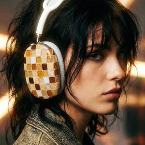 May include: White and gray headphones with earcups featuring a mosaic pattern of brown, tan, and cream-colored squares. The headphones are worn by a person with dark hair and a denim jacket.