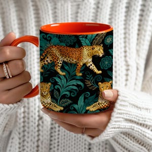Puede incluir: Taza de café con asa naranja y diseño de estampado de leopardo. La taza presenta un fondo negro con follaje verde azulado e ilustraciones de leopardos. El interior de la taza es naranja.