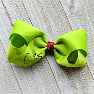 May include: A green Grinch-themed hair bow with a red glitter centre. The bow is made of grosgrain ribbon and has a printed image of the Grinch's face.