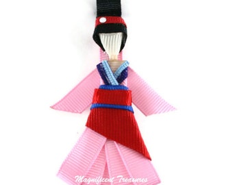 May include: A pink ribbon doll with a red and blue dress, black hair, and a black hat with a red dot. The doll is wearing a pink ribbon dress with a red and blue sash. The doll is wearing a black hat with a red dot. The doll is made of ribbon and is a hair clip.