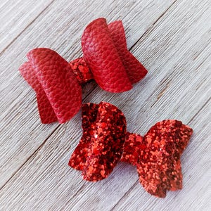 May include: Two red hair bows. One bow is made of textured red material, and the other is covered in red glitter. Both bows are on a light wood grain surface.
