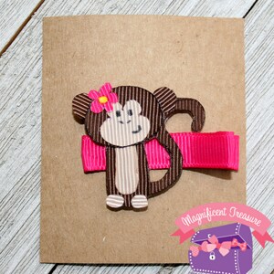 May include: A brown monkey hair bow with a pink flower and a pink ribbon. The bow is attached to a brown alligator clip.