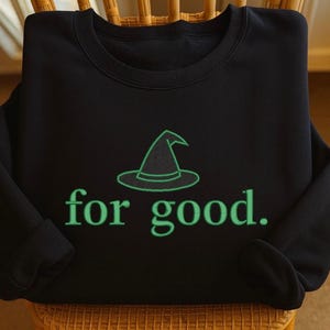 May include: Black sweatshirt featuring a green witch's hat and the words "for good." The design is centrally placed on the front of the crew neck top. The long-sleeved top is displayed on a wooden chair.