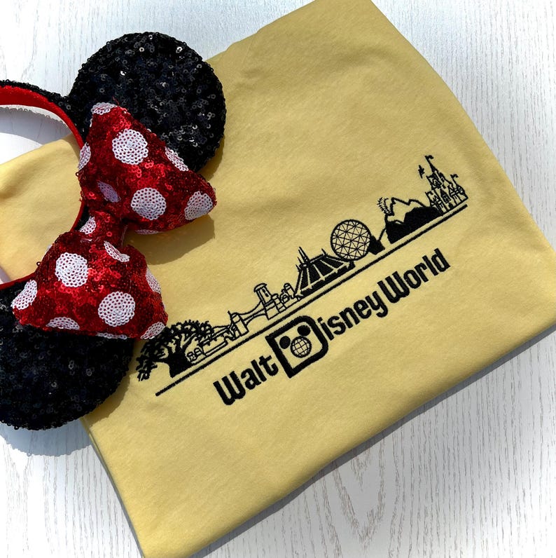 K&ouml;nnte beinhalten: Ein gelbes T-Shirt mit einer schwarzen Grafik der Skyline von Walt Disney World und Text. Neben dem Shirt befinden sich ein Paar schwarze, paillettenbesetzte Micky-Maus-Ohren mit einer rot-wei&szlig; gepunkteten Schleife.