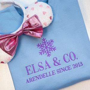 Puede incluir: Sudadera azul claro con el texto bordado "ELSA & CO. ARENDELLE SINCE 2013" y un dise&ntilde;o de copo de nieve morado. Una diadema blanca con orejas de lentejuelas y un lazo rosa se coloca sobre la sudadera.