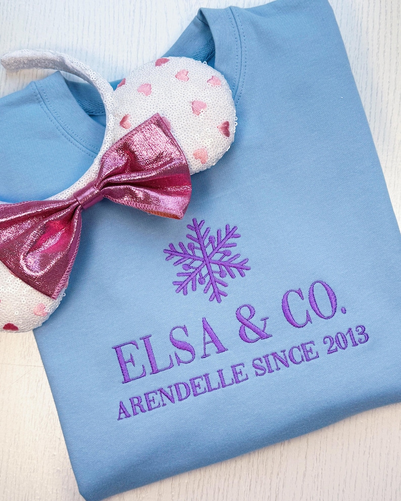 Puede incluir: Sudadera azul claro con el texto bordado "ELSA & CO. ARENDELLE SINCE 2013" y un dise&ntilde;o de copo de nieve morado. Una diadema blanca con orejas de lentejuelas y un lazo rosa se coloca sobre la sudadera.
