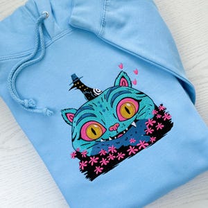 May include: Light blue hoodie with a whimsical cat face embroidered on the front. The cat has teal fur, pink eyes and ears, and a black hat. Pink flowers and hearts surround the cat's face, creating a playful design.