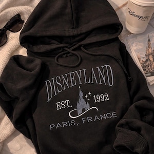 May include: A white ceramic mug with a black handle and the text "But First, Coffee" in a black, handwritten-style font. The mug is on a white surface.