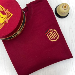 May include: A maroon crew neck sweatshirt with a gold embroidered emblem and a matching hat. The hat is round with a black band and gold text. A gold bell with black base is also present.