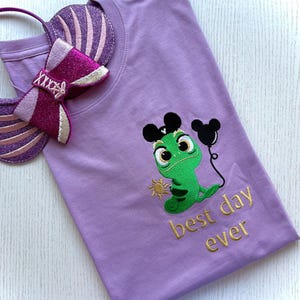 May include: A lilac t-shirt with an embroidered green chameleon wearing Mickey Mouse ears and holding a black balloon. The text 'best day ever' is embroidered in gold. A matching headband with a bow is also visible.