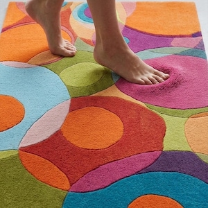 Puede incluir: Una alfombra de ba&ntilde;o rectangular con un dise&ntilde;o vibrante de c&iacute;rculos superpuestos en tonos naranja, rosa, azul, verde y morado. La alfombra de felpa mide aproximadamente 76 cm de largo y 51 cm de ancho.