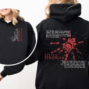 May include: Black hoodie with a hood and long sleeves. The front features a graphic of a gas mask and the text "I STRANGER". The back displays a red spider web design with a spider and the words "LITTLE STRANGER".