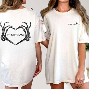 May include: Off-white t-shirt featuring a black skeleton hand heart design on the back with the text "STFUATTDLAGG". The front of the shirt has the text "GOOD GIRL" in black. The shirt has short sleeves and a relaxed fit.