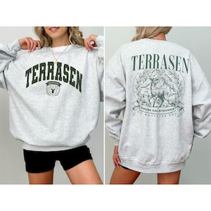 Puede incluir: Una sudadera gris claro de cuello redondo con texto verde oscuro en el frente que dice TERRASEN junto a un pequeño escudo. La parte posterior muestra un gráfico grande de un ciervo, follaje y el texto TERRASEN HOUSE GALATHYNIUS TO WHATEVER END.