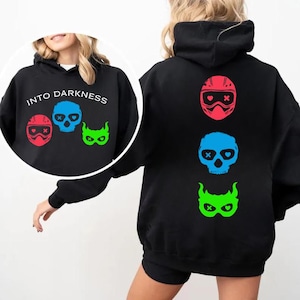 Può includere: Felpa nera con la scritta "INTO DARKNESS" e tre grafiche: un casco rosso, un teschio blu e una maschera verde. Il retro della felpa presenta le stesse tre grafiche. Un capo casual e confortevole.