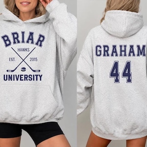 以下が含まれることがあります： 前面に「Briar University Hawks」の文字とクロスしたホッケーのスティック、年号「2015」がプリントされたライトグレーのパーカー。パーカーの背面には、名前「Graham」と数字「44」がネイビーブルーで表示されています。