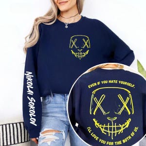 Op de afbeelding: Marineblauwe sweatshirt met een neongele afbeelding van een lachend masker op de voor- en achterkant. De linkermouw heeft de naam "Nikolai Sokolov" in het wit. De achterkant van de sweatshirt heeft de tekst "Even if you hate yourself, I'll love you for the both of us."